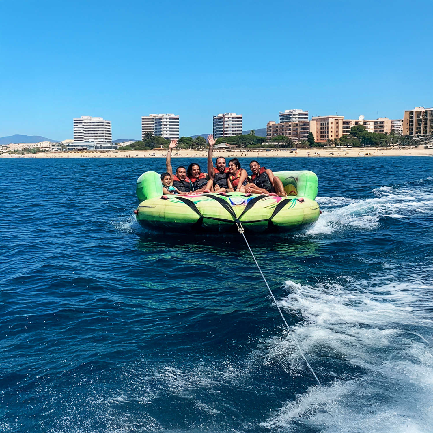 People on the Crazy Sofa - Inflatable Towable being pulled by a boat in the ocean with buildings in the background.